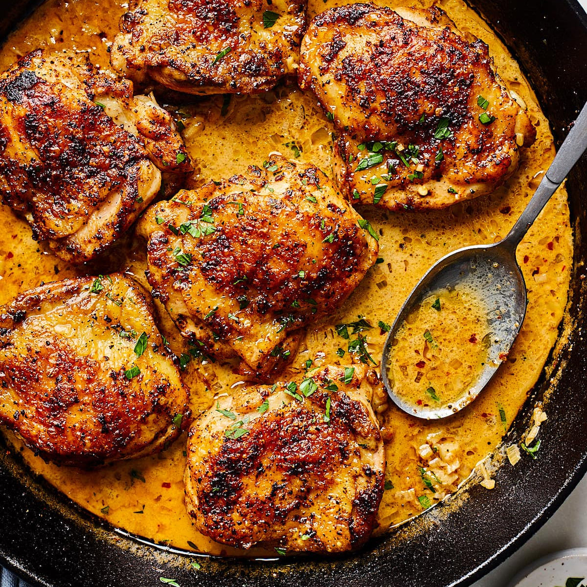 Overhead close-up of six seasoned chicken thighs in a creamy orange-yellow sauce within a black cast-iron skillet, with a metal spoon and fresh parsley garnish.